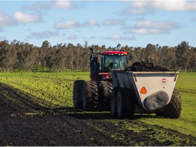 Tractor applying biosolids using spreader in field with trees and sky in background